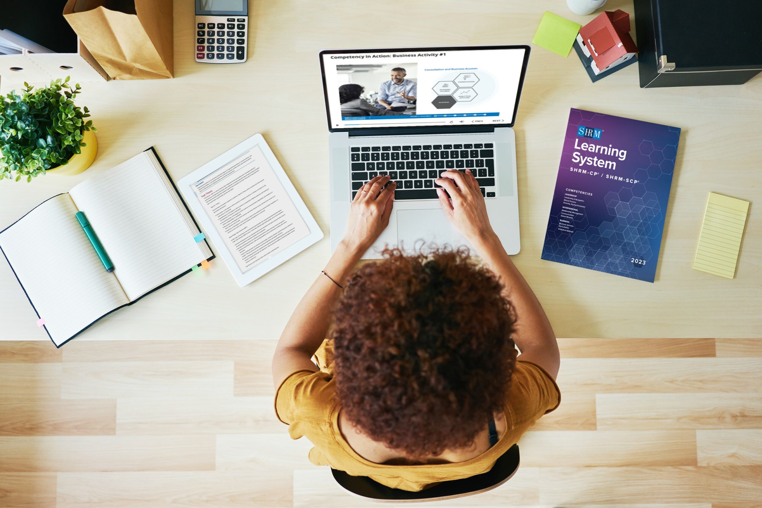 Woman sits at computer with SHRM workbook on desk next to her.